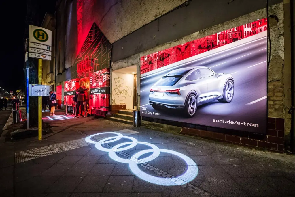 A nighttime street scene featuring a bright billboard advertising an Audi e-tron car, with the illuminated Audi logo projected onto the pavement in front of the display. BattleRoyalStudios