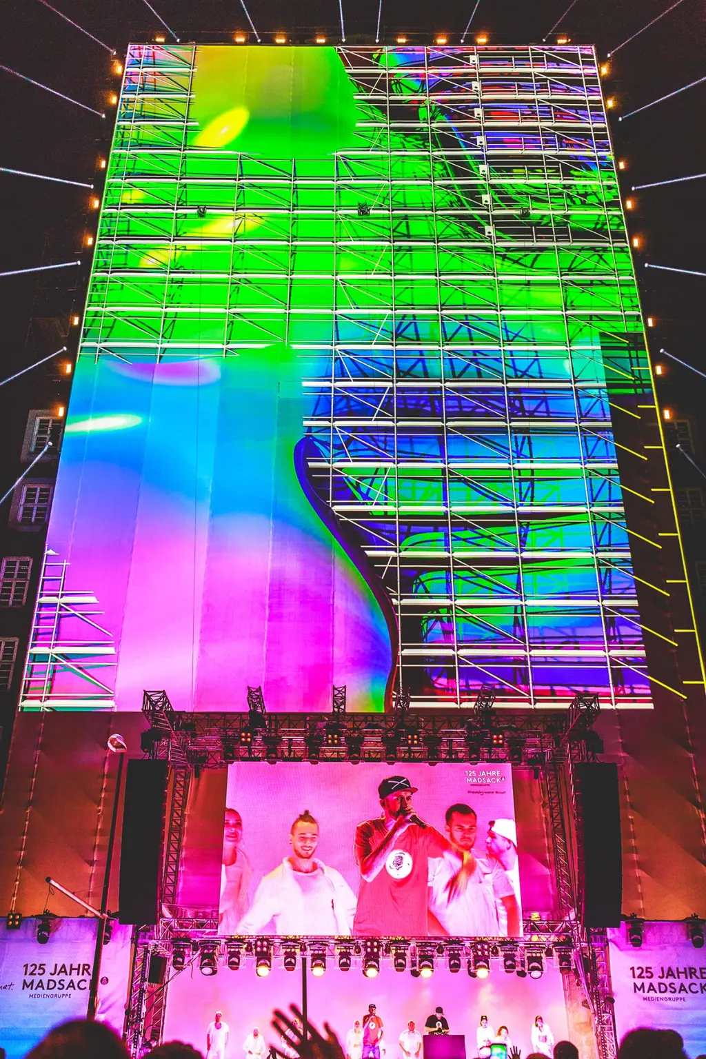 Giant LED screen displaying vibrant colors and abstract visuals during a live performance with a rapper in a red shirt on stage. The event is celebrating "125 Jahre Madsack Mediengruppe." Battle Royal Studios