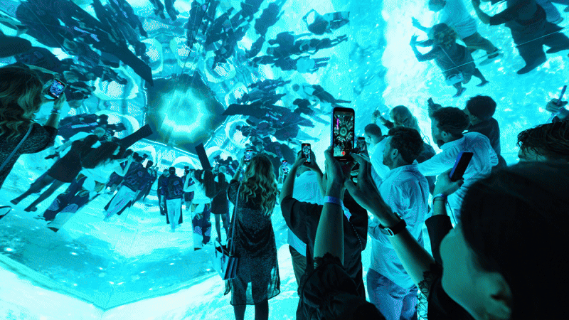 A person poses playfully with glowing lights, while another takes a photo using a smartphone, surrounded by hanging strands of illuminated lights. Battle Royal Studios