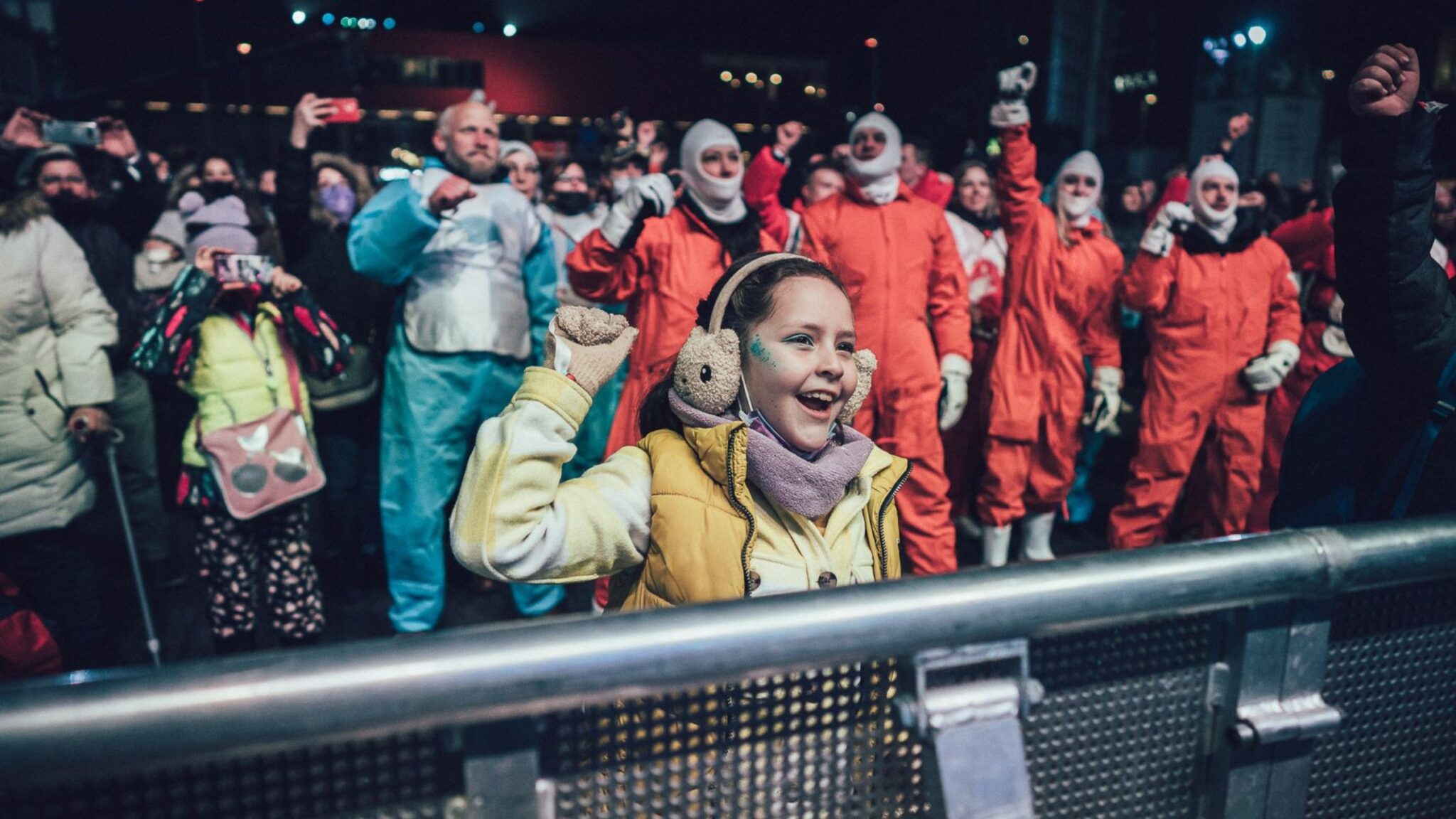 Young girl in earmuffs smiling and raising her fist in excitement, with a group of people in colorful protective suits in the background. Battle Royal Studios.