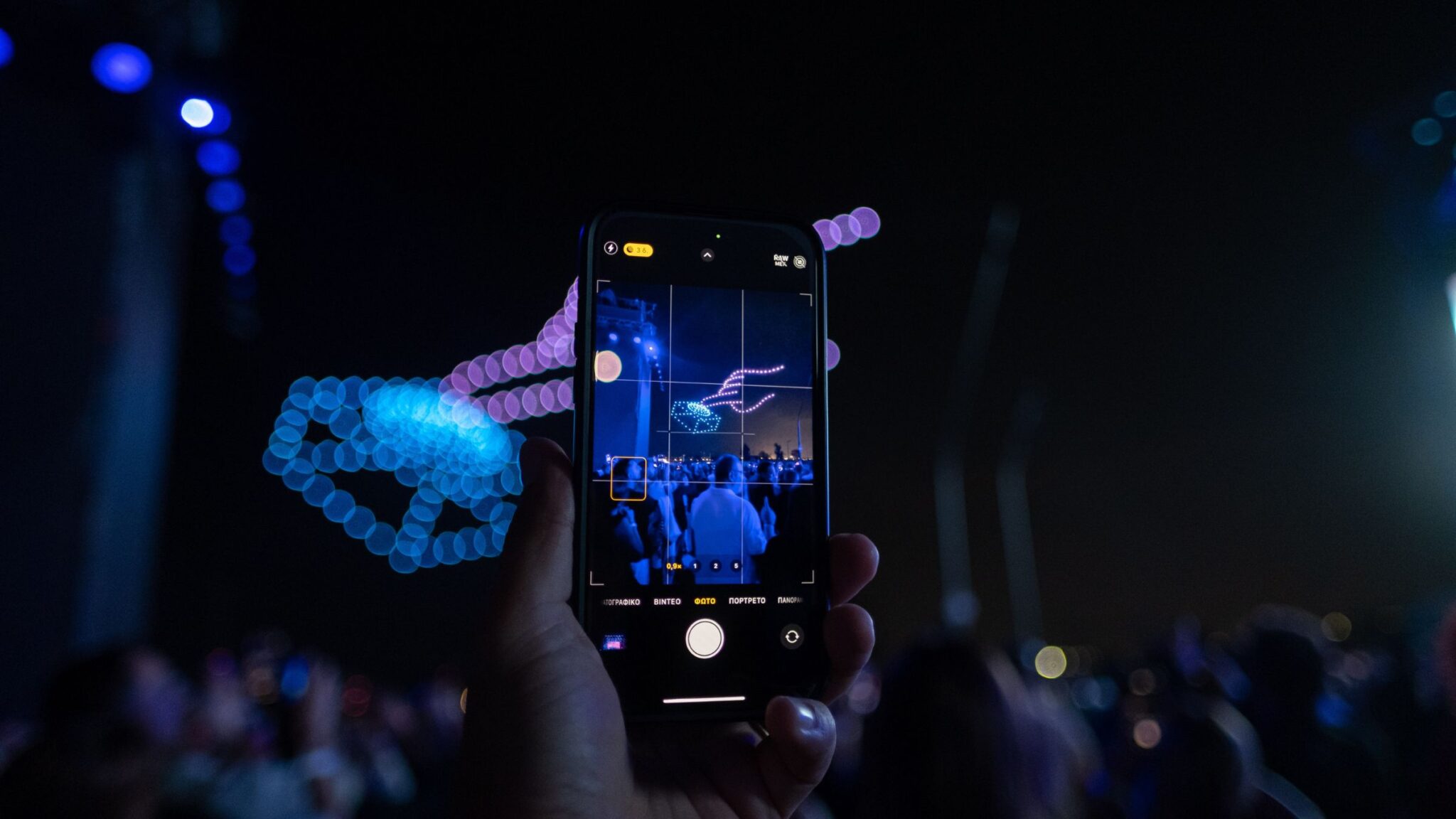 A person holds up a phone, capturing a light display of drone formations in the night sky, with blue lights creating a shape. Battle Royal Studios