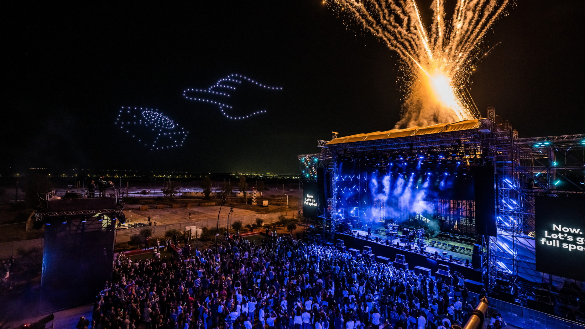 A large crowd enjoys a nighttime concert with fireworks and drones creating light patterns above the stage. Battle Royal Studios
