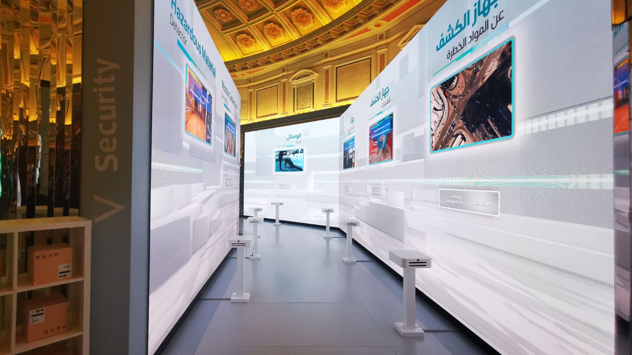 A modern exhibition space with large digital screens showcasing information about hazardous material detection and other security technologies, under an ornately designed ceiling. Battle Royal Studios