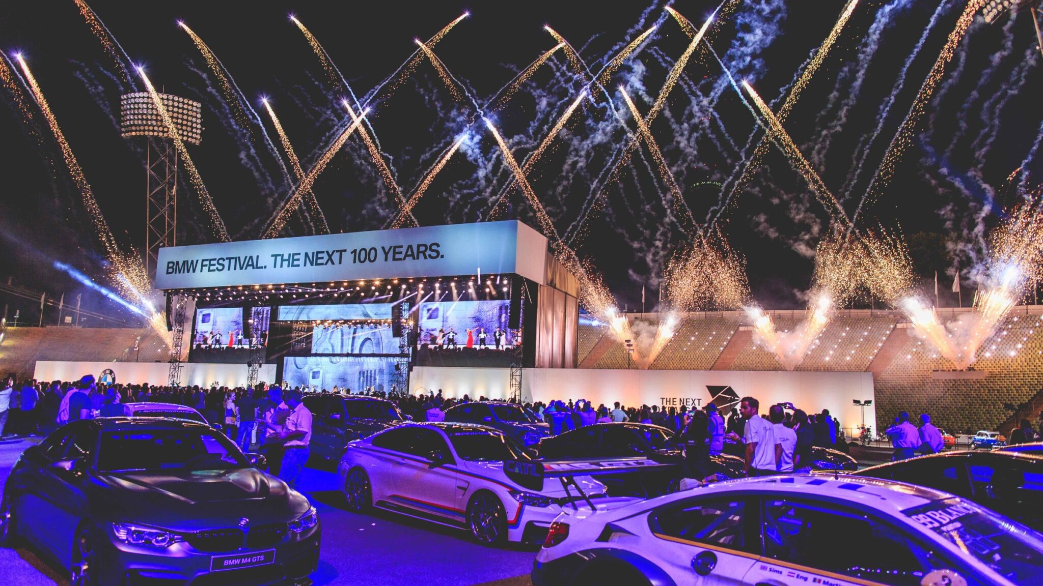 A large crowd gathers at the BMW Festival with cars displayed in the foreground and fireworks lighting up the night sky above the event stage. Battle Royal Studios