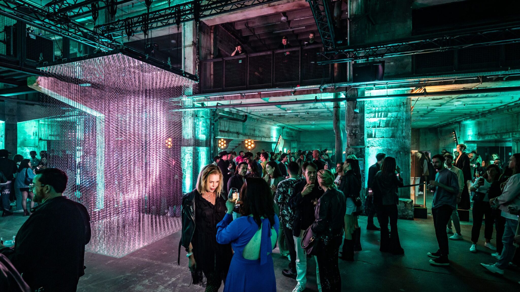 People socializing in an industrial-style space with green lighting, featuring a hanging installation of small lights and a modern atmosphere. Battle Royal Studios.