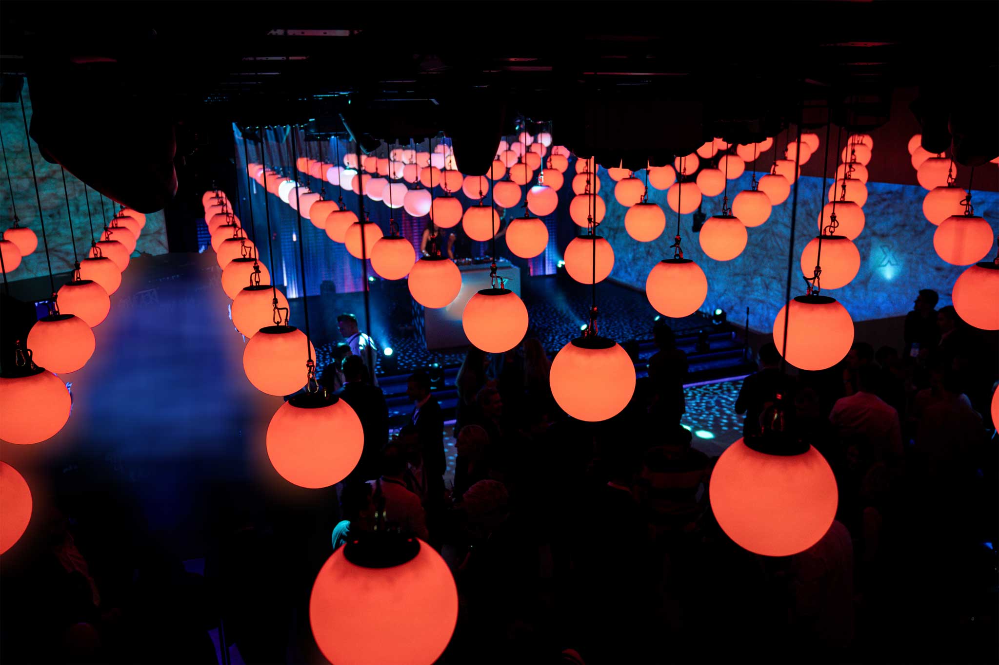 A ceiling installation of glowing red orb lights illuminating a dimly lit event space with guests and a DJ in the background Battle Royal Studios