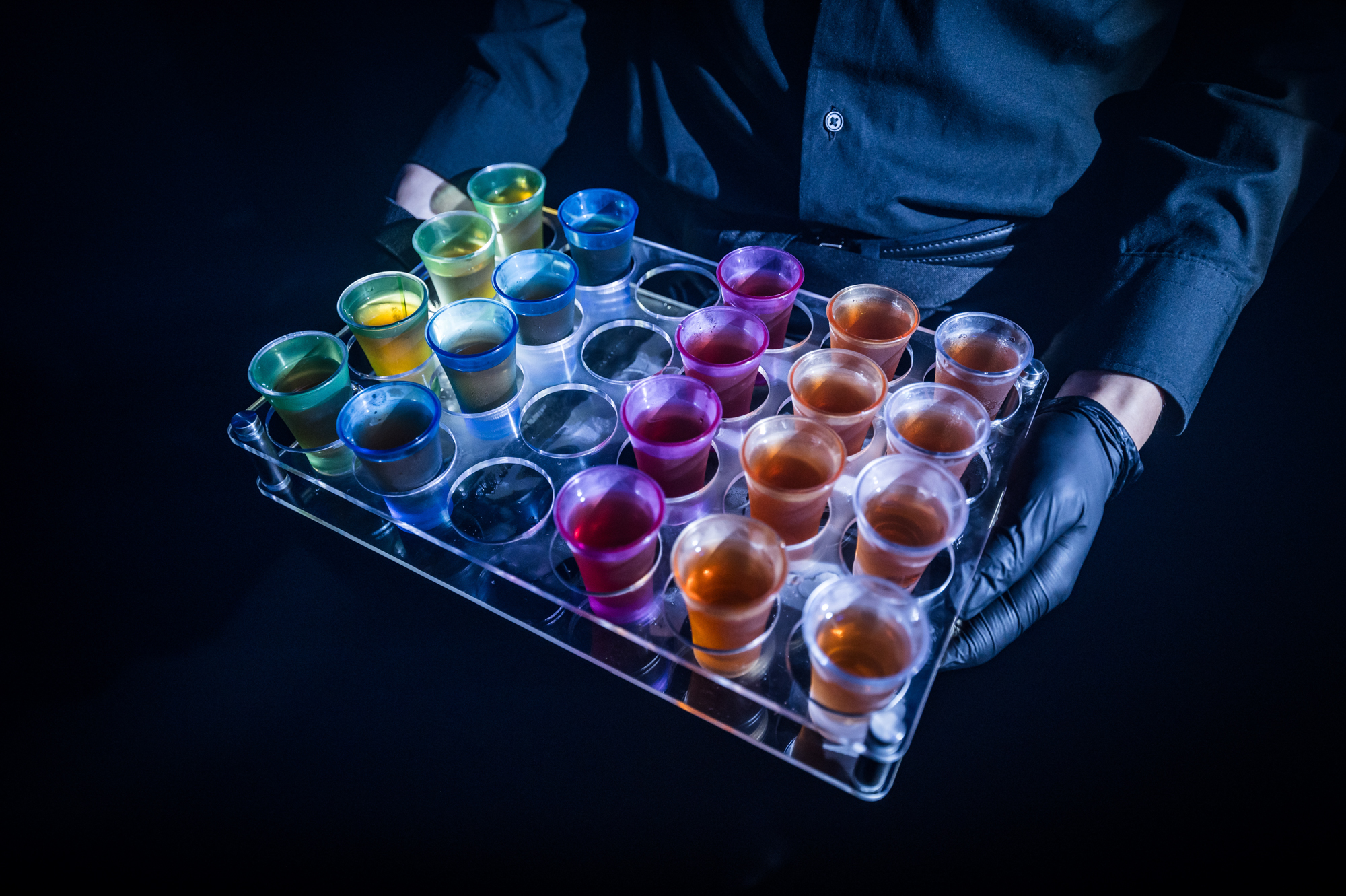 A server in black gloves presents a tray of colorful illuminated drinks in vibrant shot glasses Battle Royal Studios